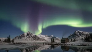 Green and purple aurora borealis dancing over snow mountains and wooden cabin in Iceland reflecting in still lake, representing Iceland as top winter destination 2026