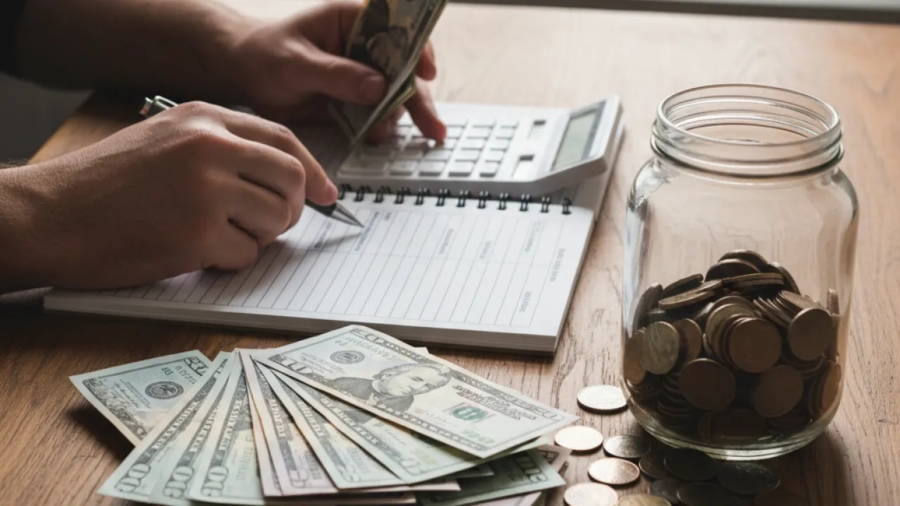 Person counting cash with calculator and savings jar on wooden table representing how to save money fast 2026